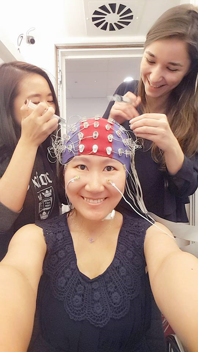 A photograph of three women. The woman in the middle is seated and is wearing a blue-and-red elastic EEG cap interspersed with white electrodes. The two other women are standing to her sides and holding transparent plastic, tube-shaped gel applicators.